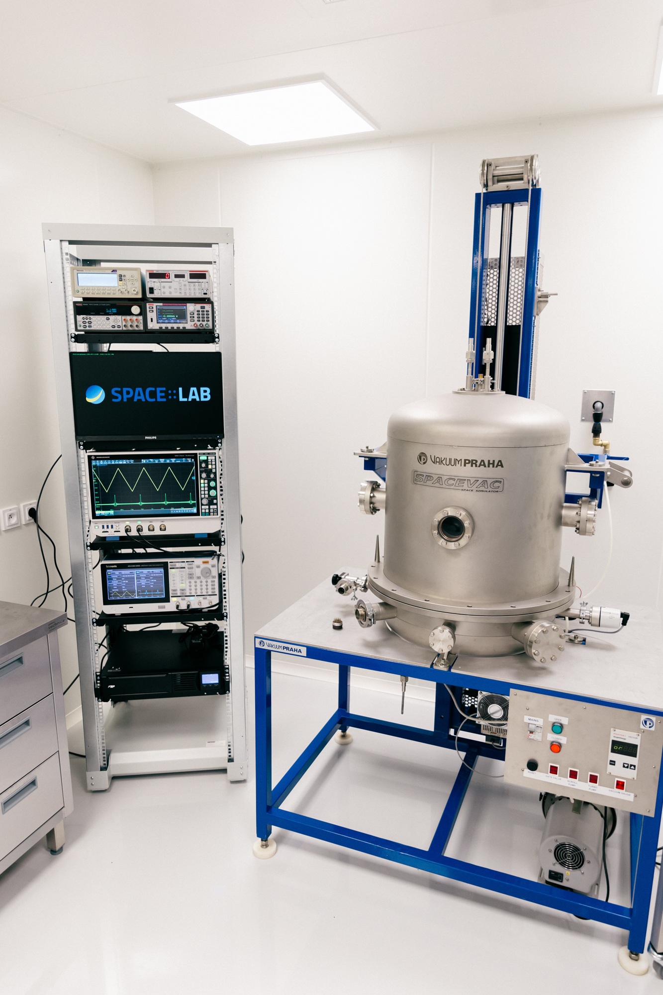 A stainless steel thermal-vacuum chamber on a blue frame beside a rack of laboratory instruments including an oscilloscope and multimeters.