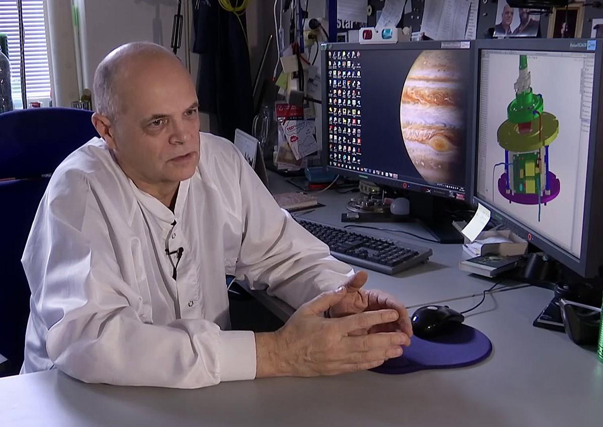 Ján Baláž seated at a desk with monitors showing Jupiter and a CAD model of the instrument.