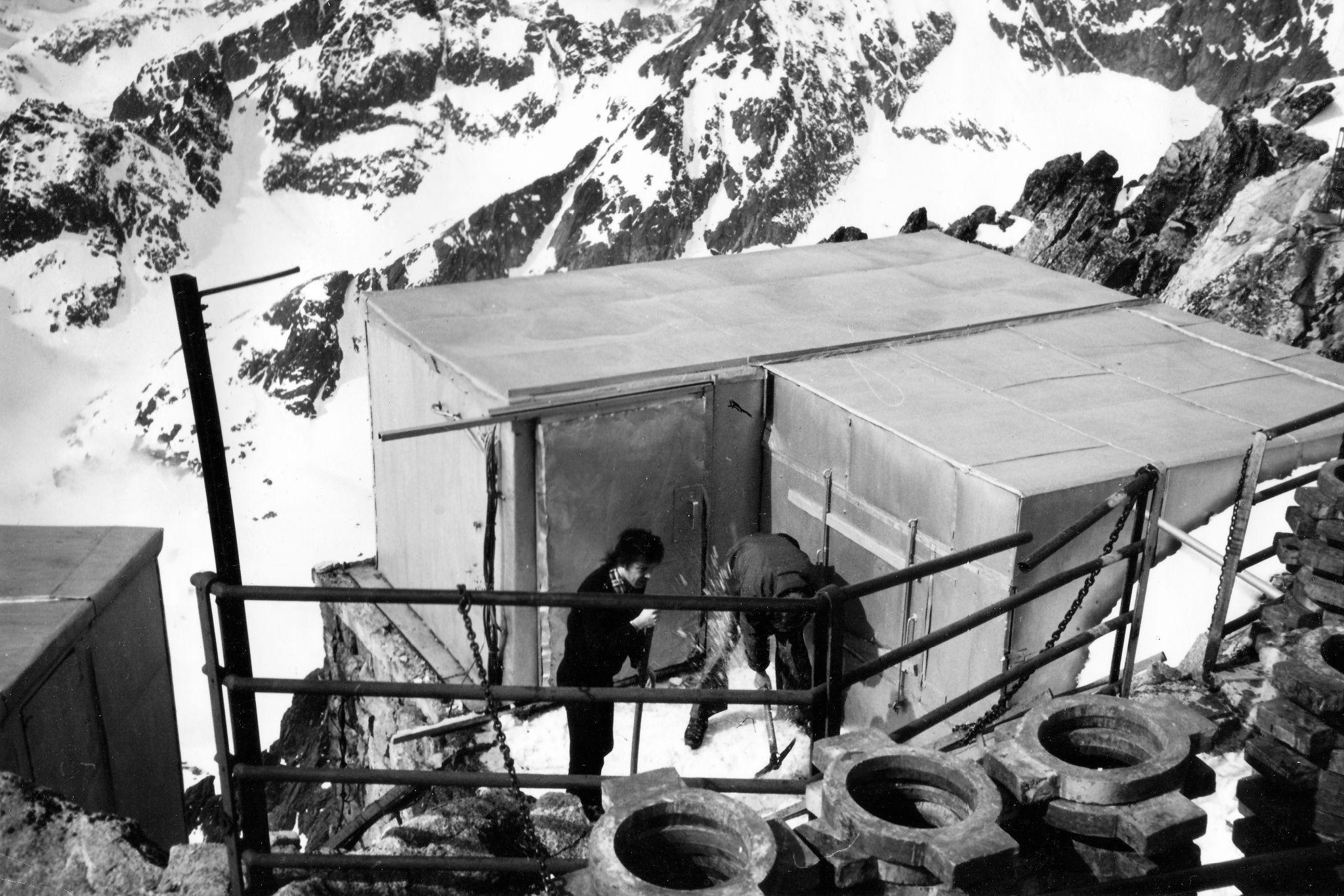 A black and white photograph of a small flat-roofed hut on a mountain summit, held down with car tyres, with two people working in front of it.
