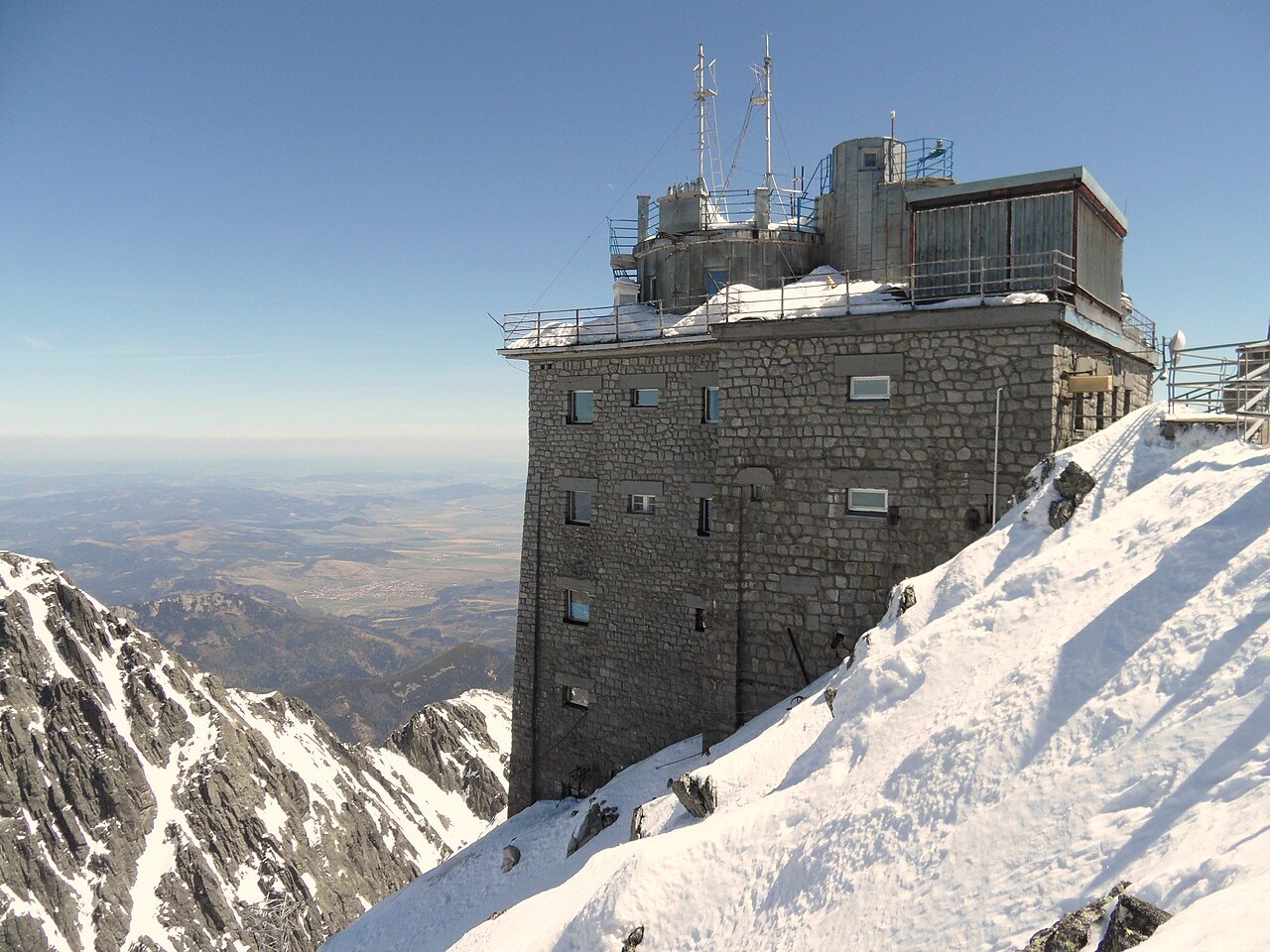 The stone observatory building on a snow-covered summit ridge, with a metal-clad measurement building standing on its roof, the Slovak lowlands visible far below.